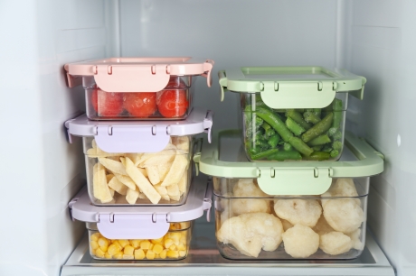 Photo of a fridge shelf with two piles of five clear food containers with lids. The containers hold cherry tomatoes, green beans, fettuccine, sweet corn, and potatoes.