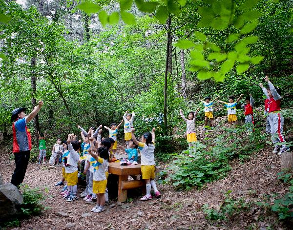 A female teacher demonstrates a power pose to her classroom of young children in a clearing in a forest. The teacher is raising her arms and the students mimic her actions.